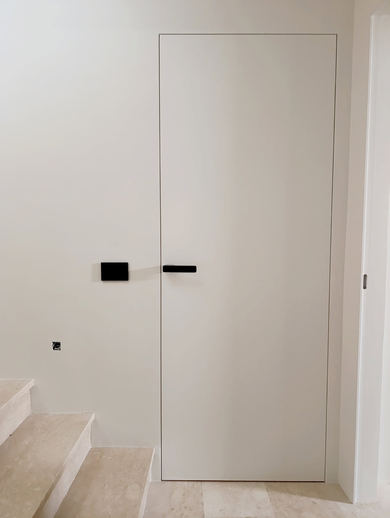 Porta a filo muro con maniglia nera accanto a una scala in pietra chiara